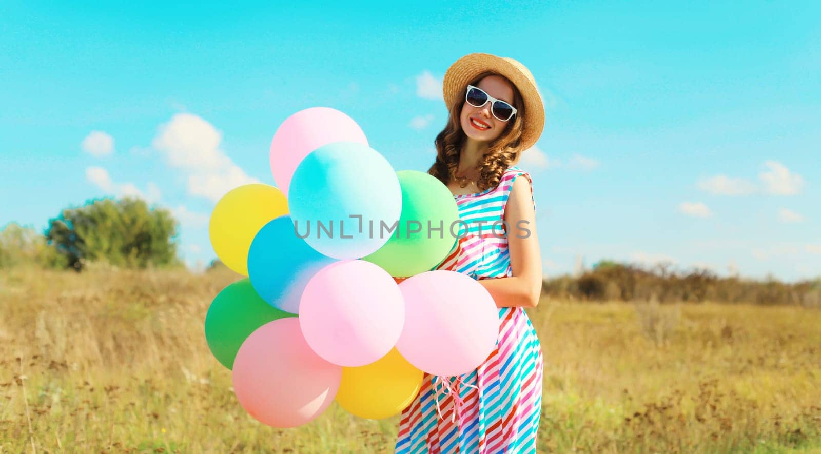 Happy smiling young woman with bunch of colorful balloons in summer straw hat on field on blue sky background
