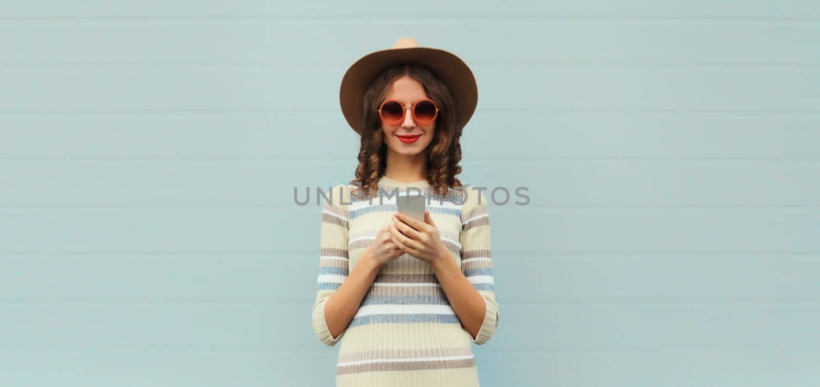 Portrait of stylish young woman with smartphone in round hat, sunglasses on gray background