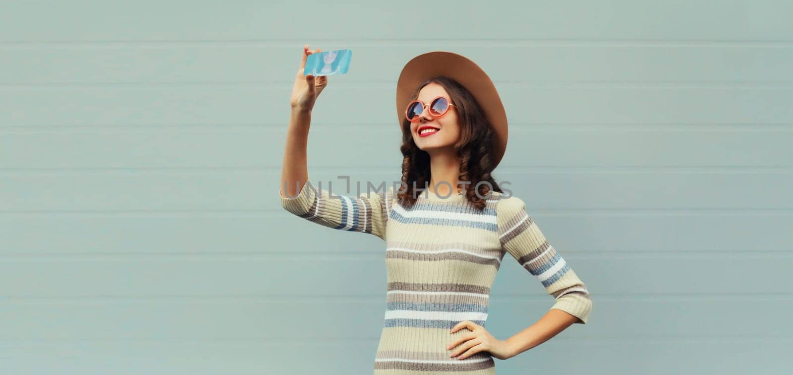 Portrait of stylish happy smiling young woman taking selfie with mobile phone in round hat, sunglasses on gray background