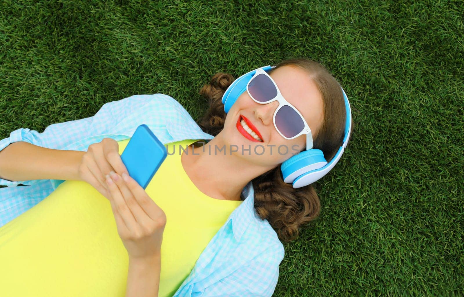 Happy young woman holding phone listening to music in headphones while lying on grass in summer park