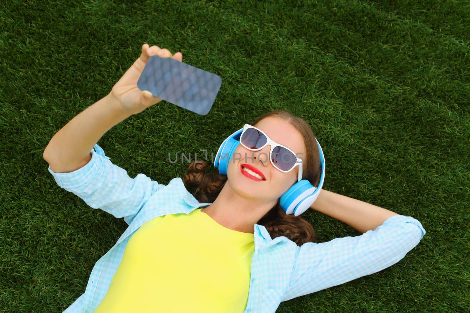 Happy young woman taking selfie with smartphone listening to music in headphones while lying on grass in summer park