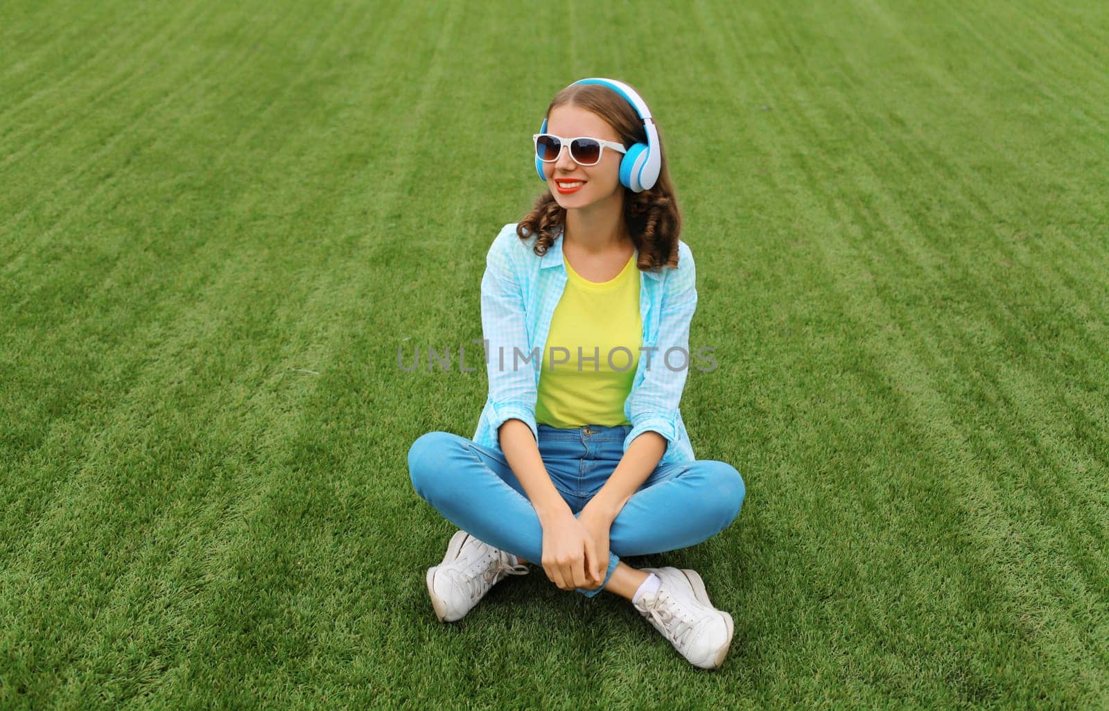 Portrait of happy smiling young woman listening to music in headphones while lying on grass in summer park
