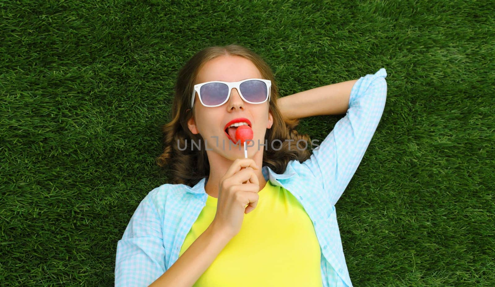 Portrait of happy teenager girl resting with lollipop lying on green grass in summer park, top view