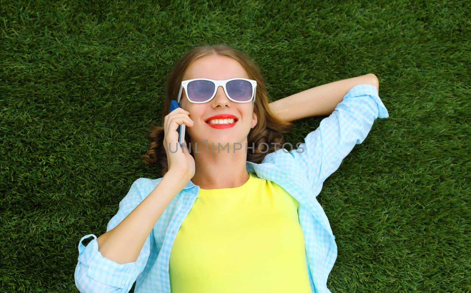 Portrait of happy smiling young woman calling on mobile phone while lying on green grass in summer park, top view