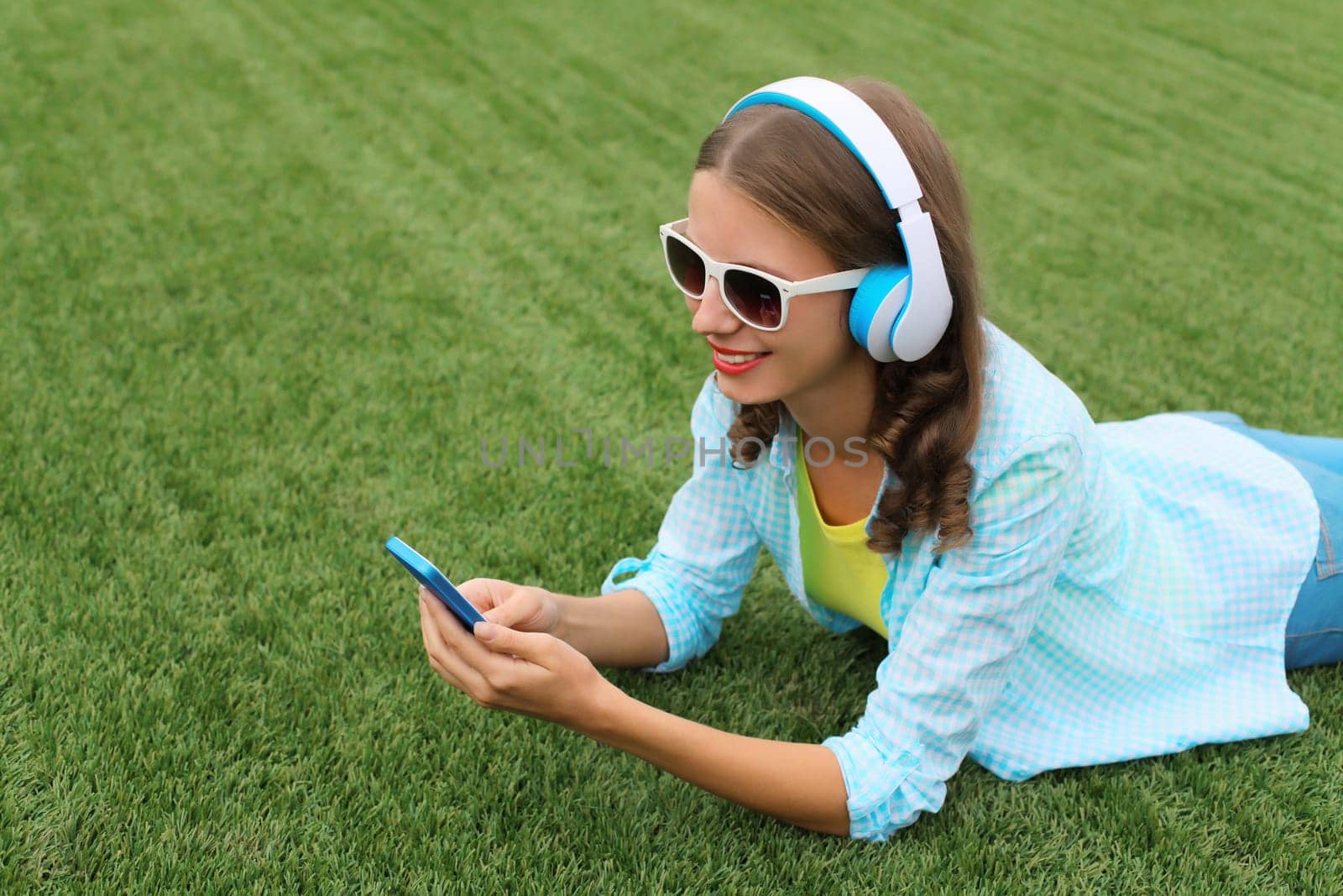 Happy young woman holding phone listening to music in headphones while lying on grass in summer park