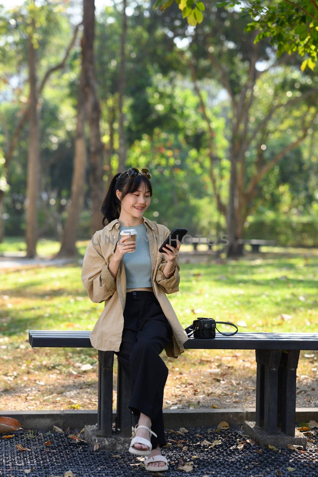Full length portrait of young woman drinking coffee and using smartphone on bench in park by prathanchorruangsak