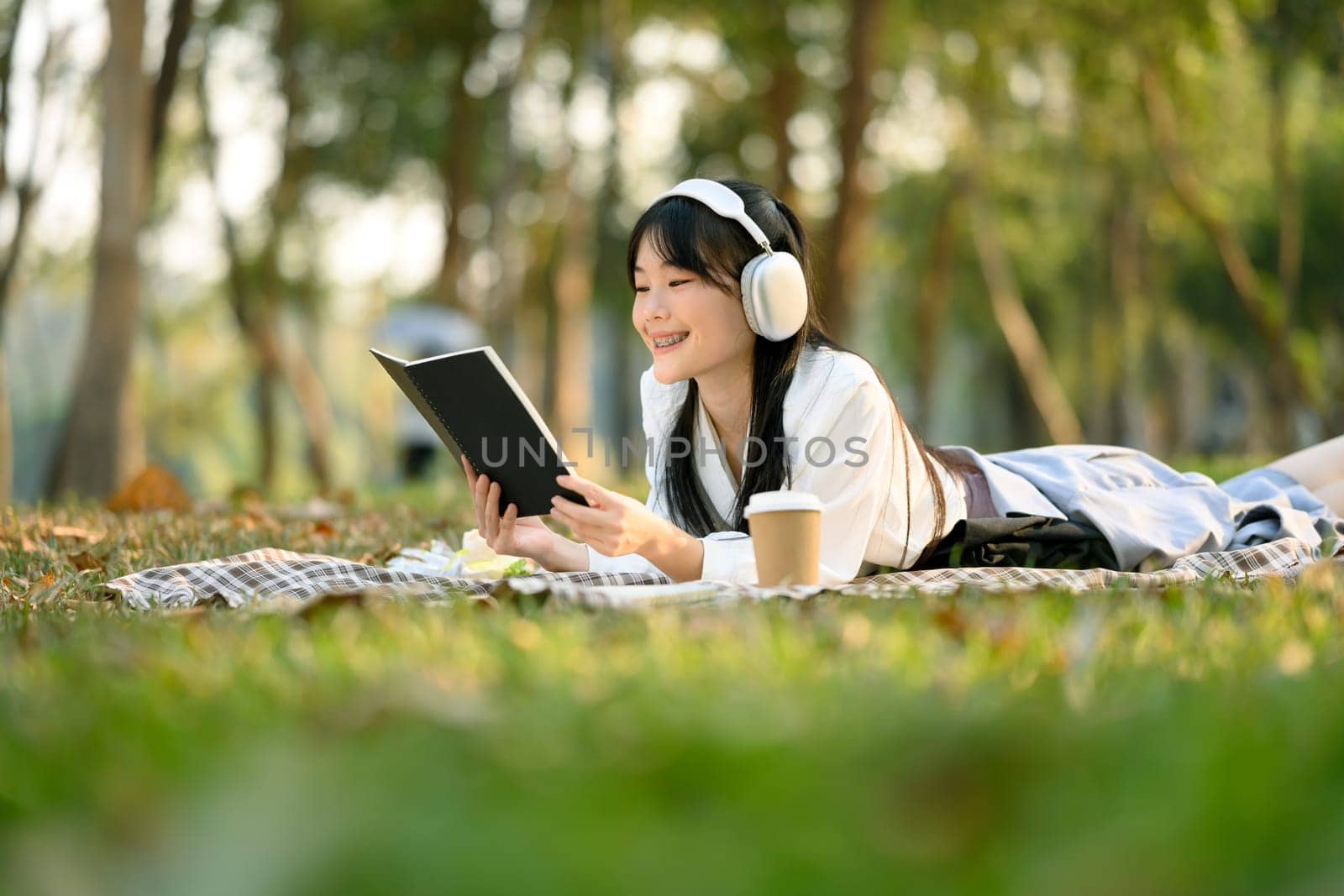 Happy young woman listening to music and reading book on picnic blanket.