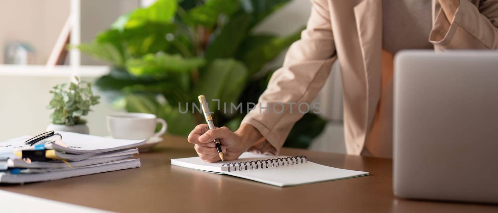 Close up of hands with pen of young businesswoman is sitting at table with laptop while taking notes, analyzing information.