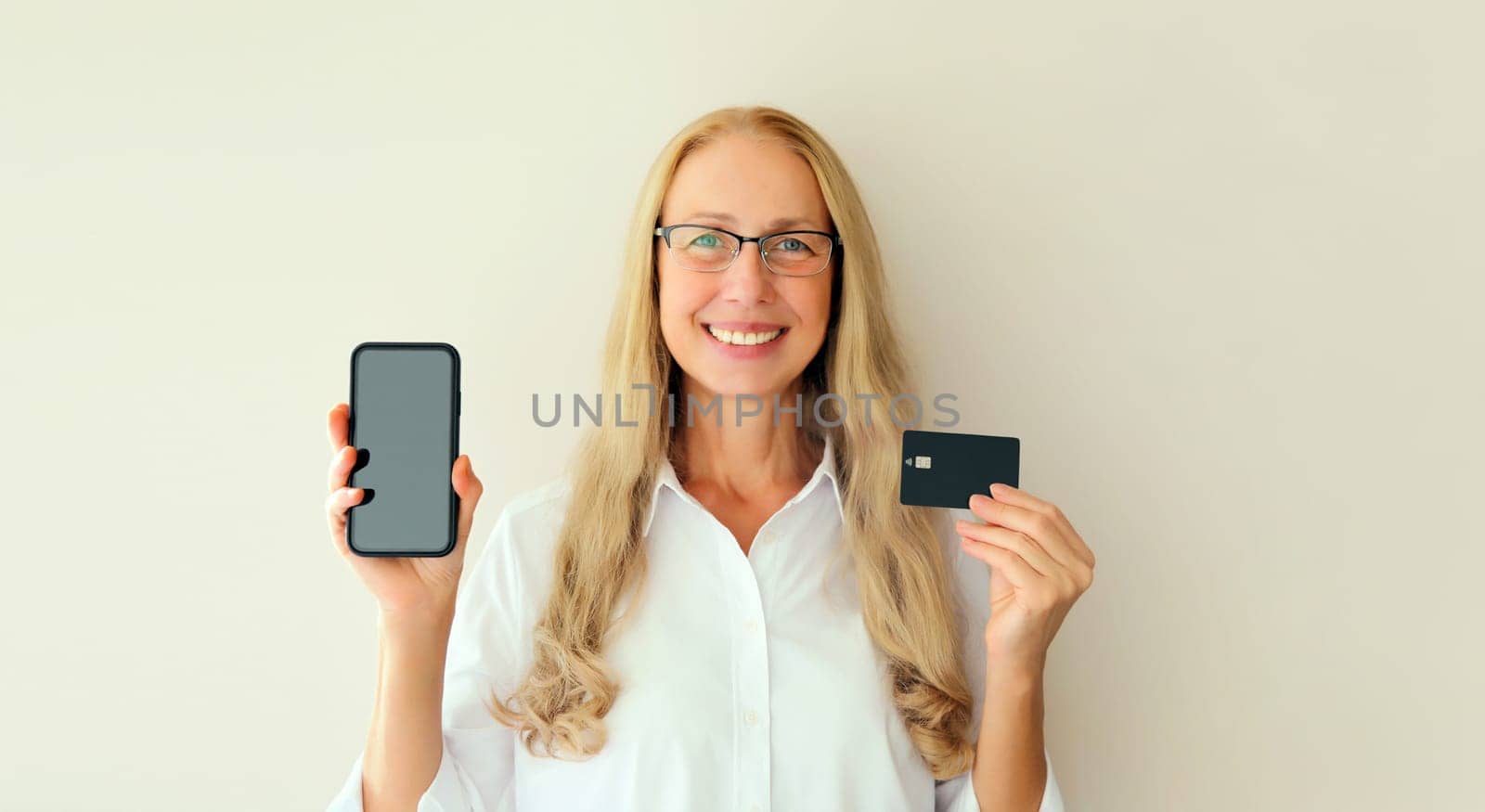 Portrait of happy smiling middle-aged woman holding plastic credit bank card and blank screen phone on white studio background