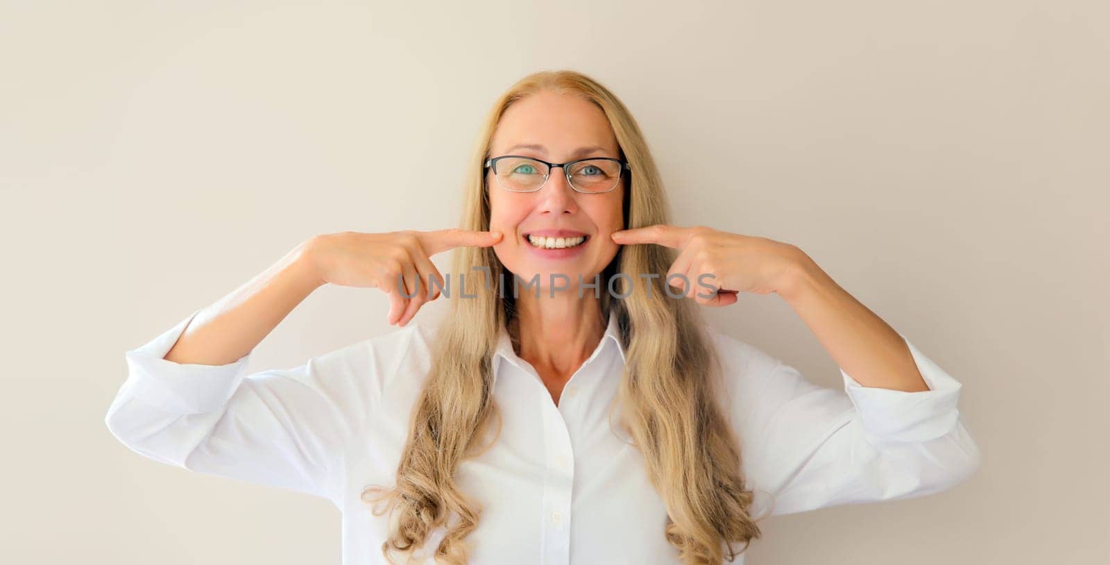 Portrait of happy smiling middle-aged woman points with fingers to her white clean teeth in eyeglasses on white studio background