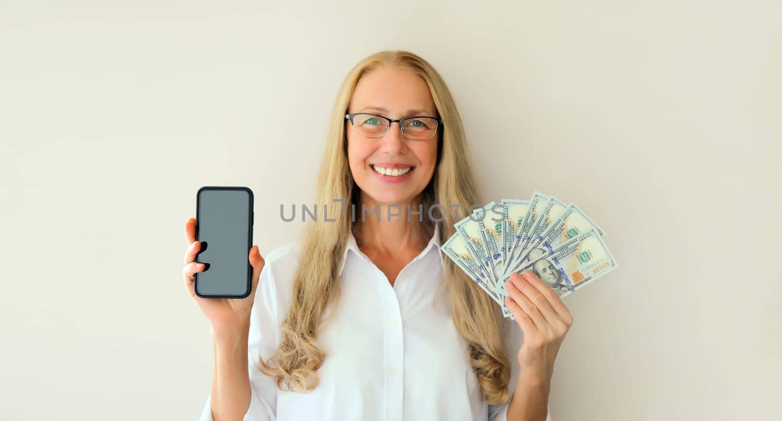 Portrait of happy smiling middle-aged woman holding blank screen phone and cash money in dollar bills on white studio background