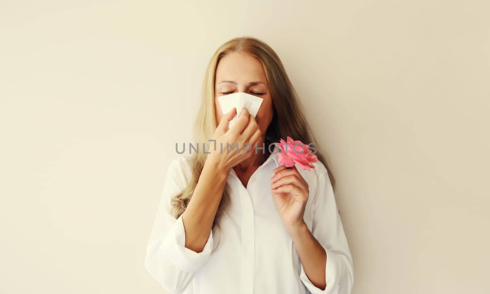 Sick exhausted woman suffering from pollen holds flower sneezing blow nose using tissue on white studio background