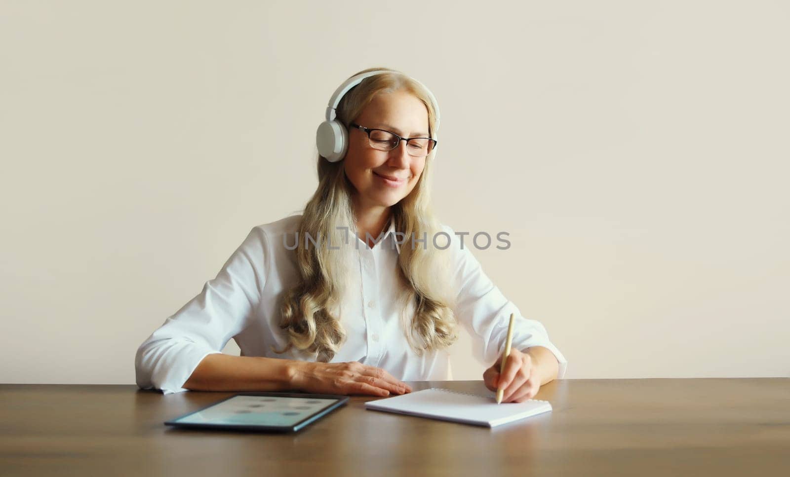 Happy relaxing middle aged woman listening to music in headphones working with digital tablet computer sitting at desk at home