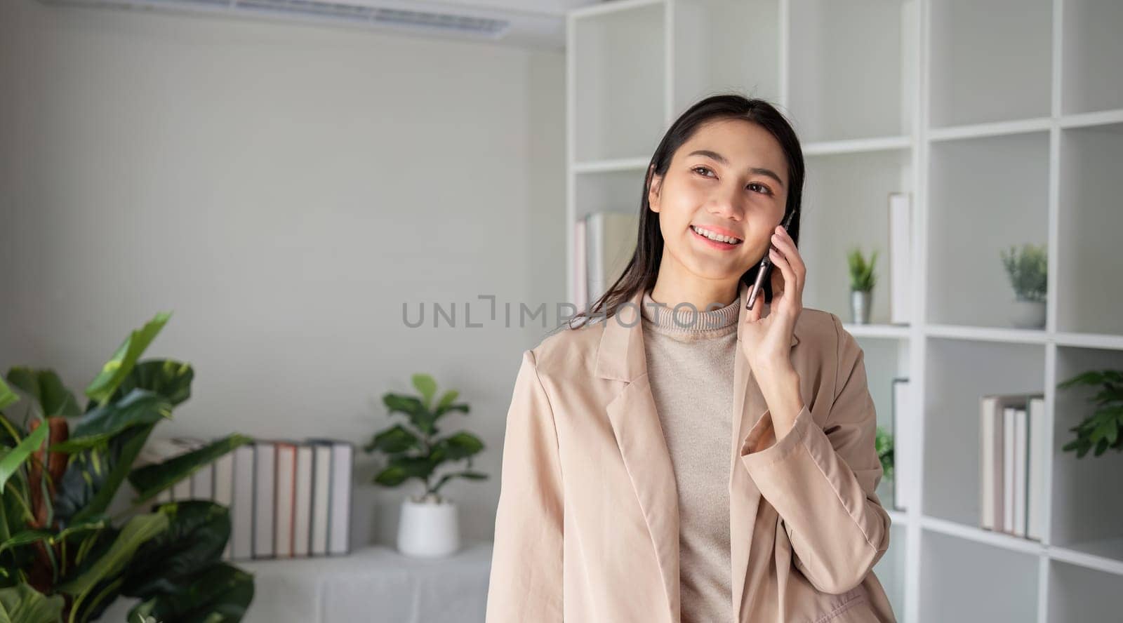 Asian businesswoman talking on the phone in an online business meeting in a modern home office decorated with lush green plants..