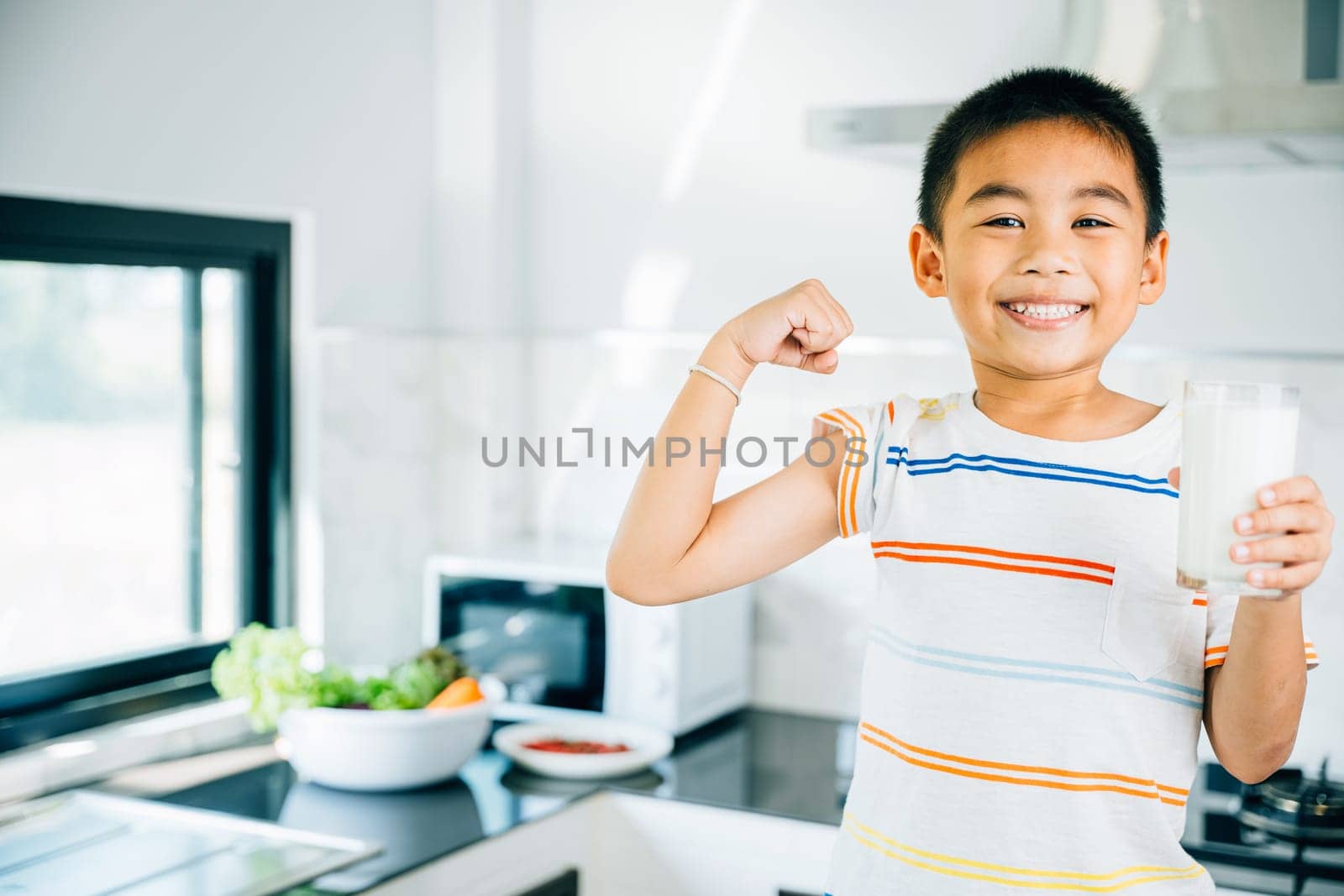Happy Asian little boy in kitchen holds milk cup, smiling. Portrait of cute son enjoying the drink. Joyful child sips calcium-rich liquid, radiating happiness at home give me.