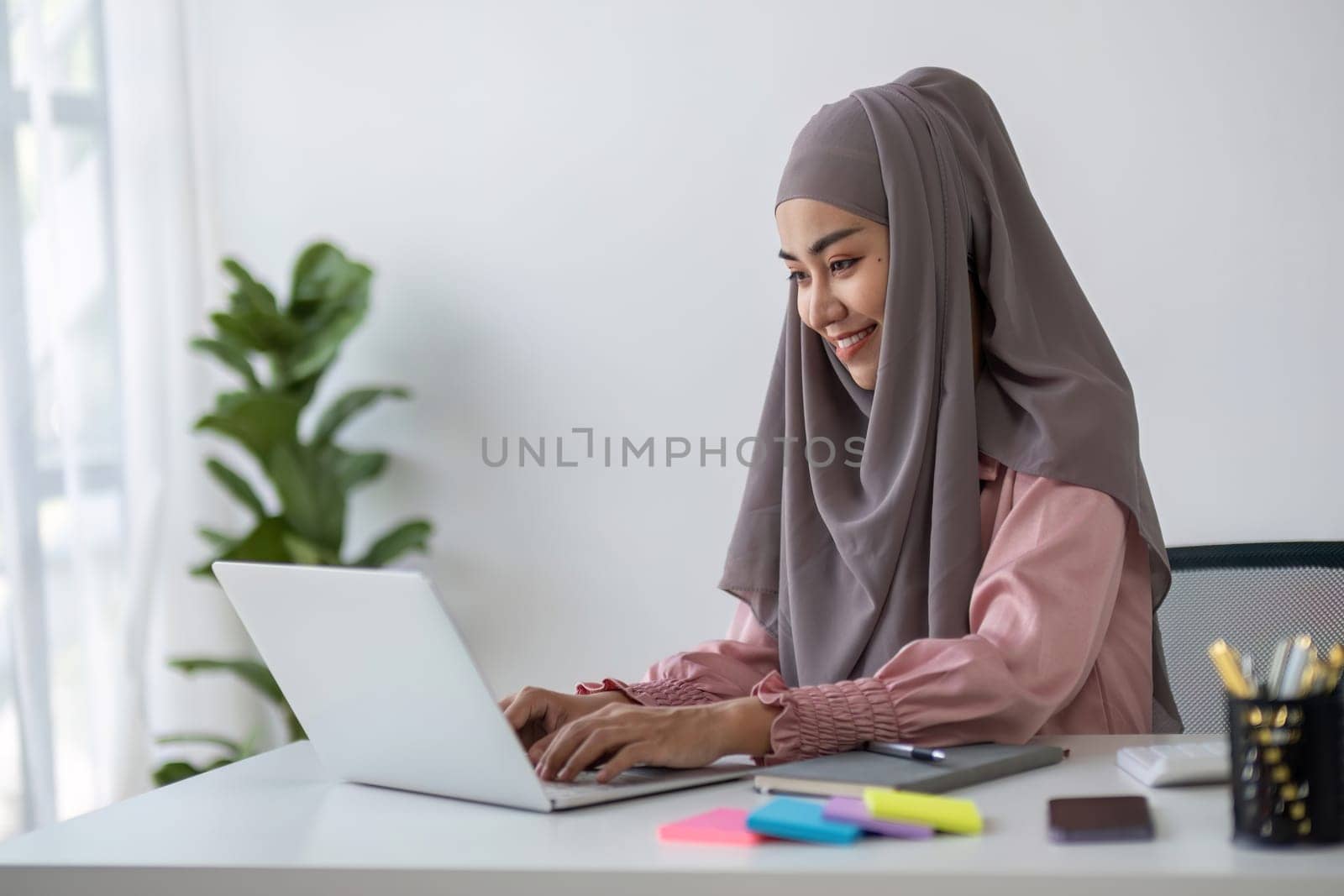 Smiling Muslim business woman Wearing a hijab while chatting online on a work desk in the office. by wichayada