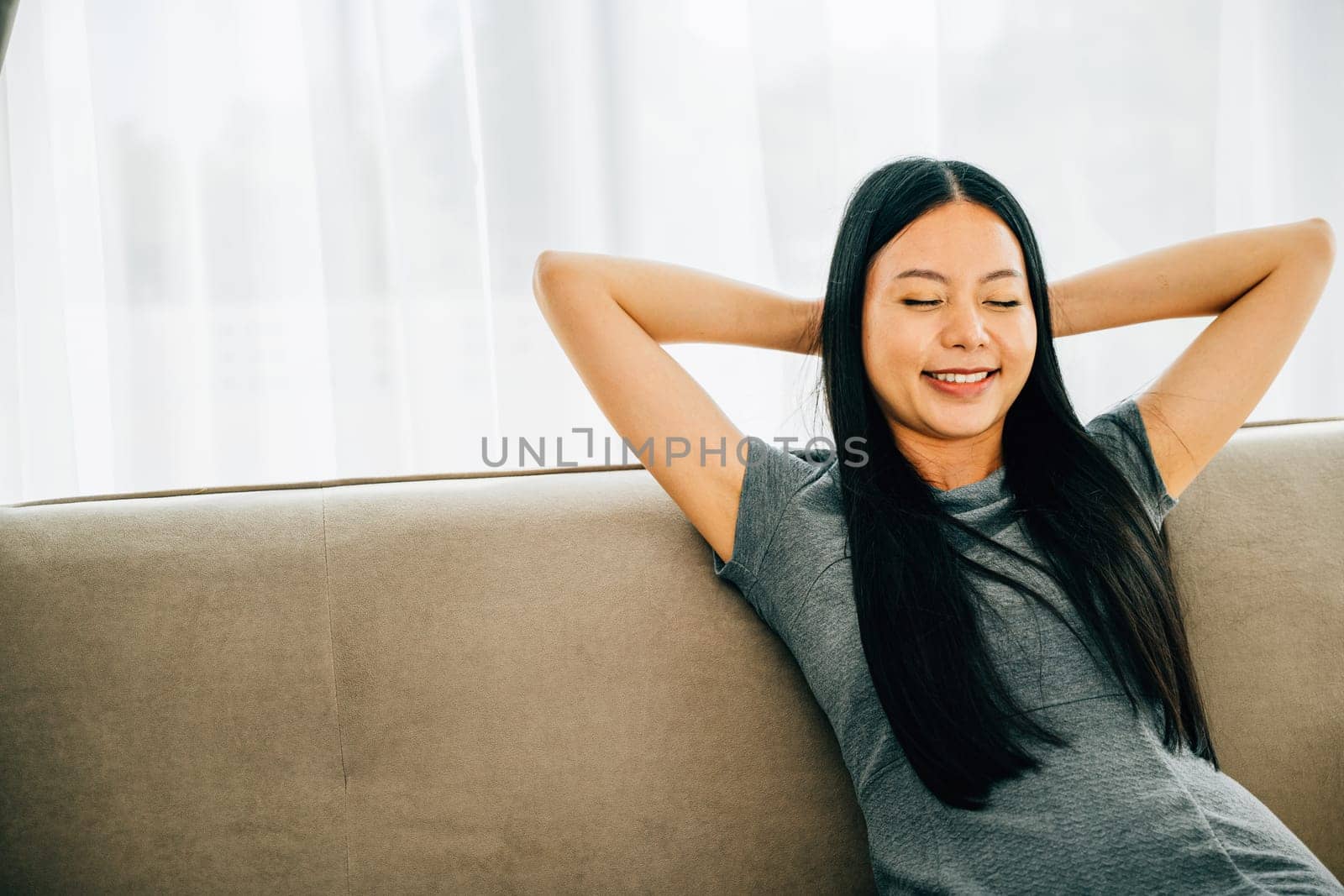 Serene woman on sofa hands behind head embraces relaxation. Dreaming and finding balance in a cozy living room. Mindfulness wellbeing and comfort at home. Kick back and relax concept