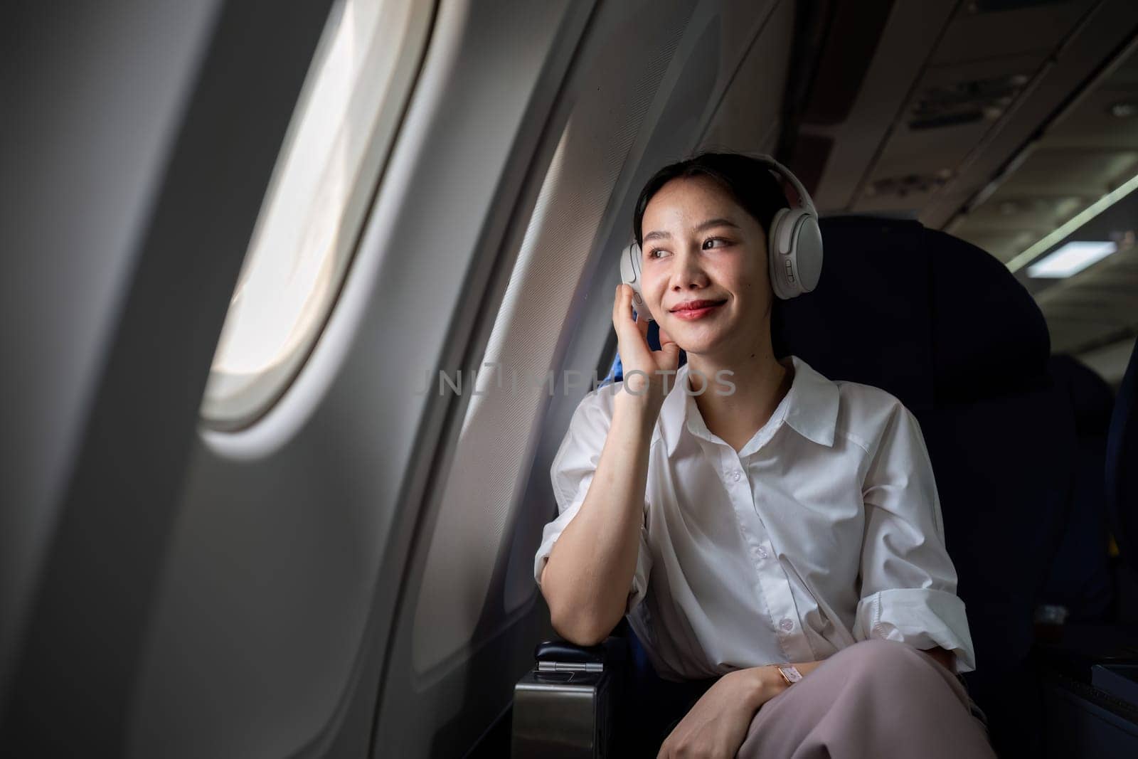 Young woman wearing headphones listening to music during travel, sitting near window in first class on airplane during flight, travel and business concept by wichayada