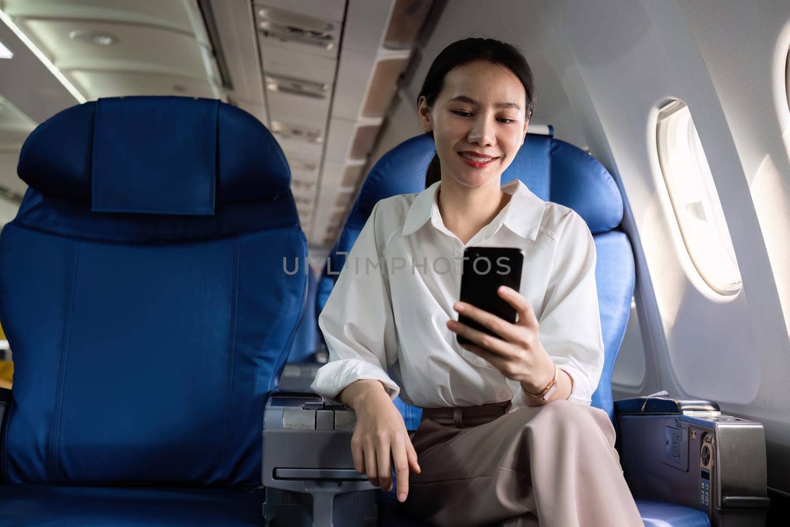 Young Asian woman checks business news on mobile phone, sitting near window in first class on airplane during flight, travel and business concept by wichayada