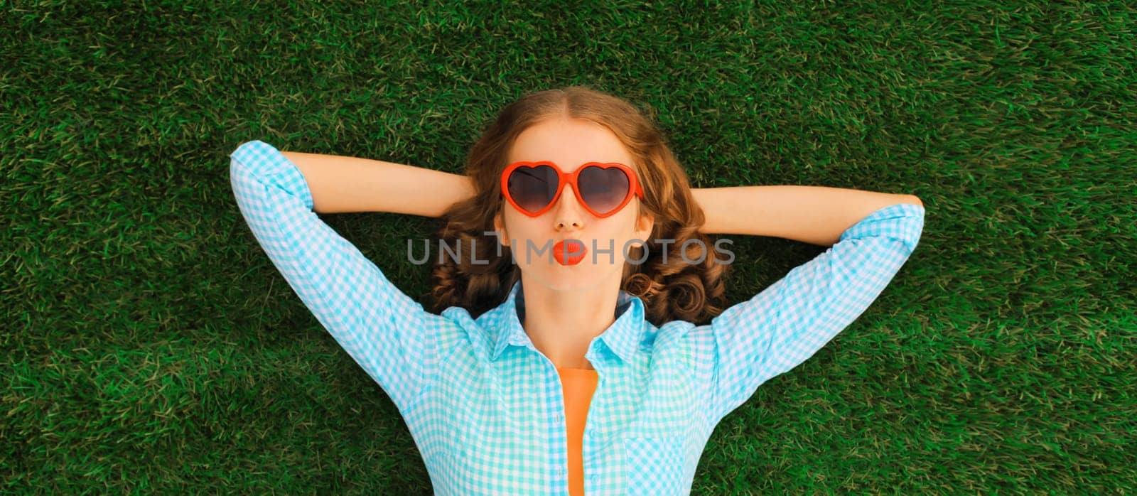 Summer portrait of happy young relaxing woman in red heart shaped sunglasses blowing kiss lying on green grass in the park