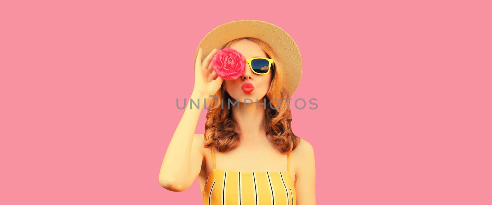 Summer portrait of stylish young woman model blowing kiss with flower buds looking for something in round straw hat on pink studio background