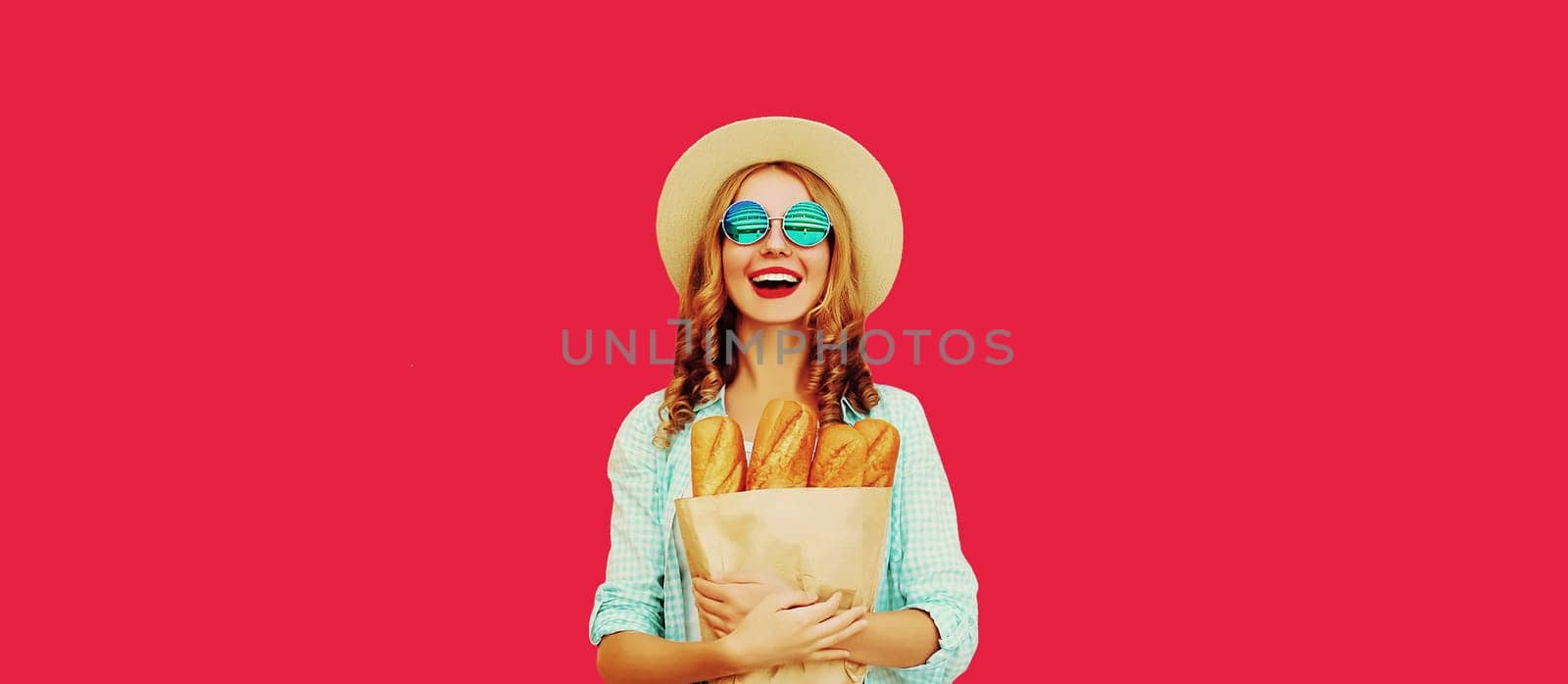 Portrait of happy cheerful smiling young woman holding grocery shopping paper bag with fresh long white bread baguette on red studio background