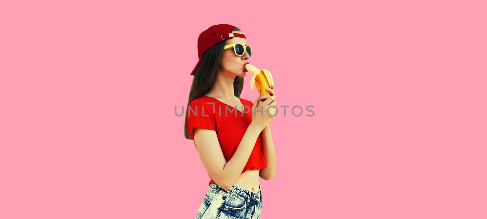 Stylish caucasian young woman eating banana in red baseball cap on pink studio background