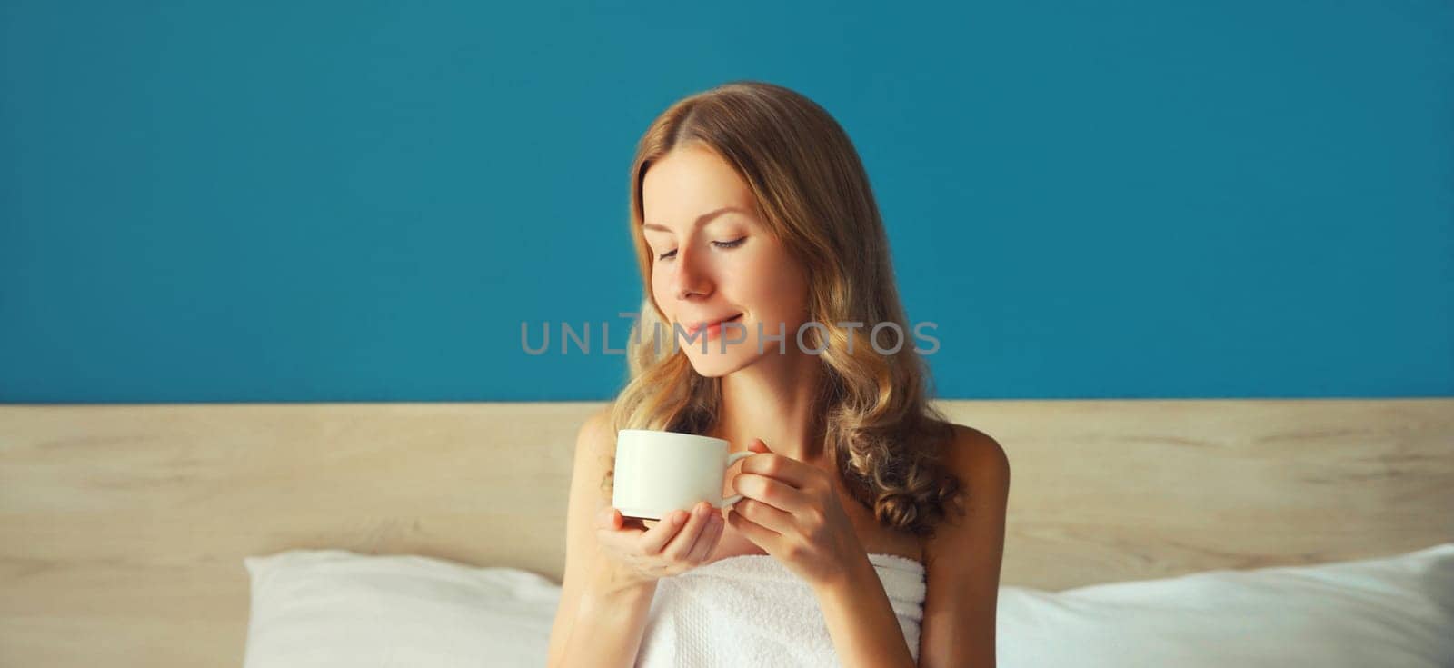 Portrait of happy satisfied young woman enjoying hot tasty cup of coffee or tea in early morning at home sitting on the bed