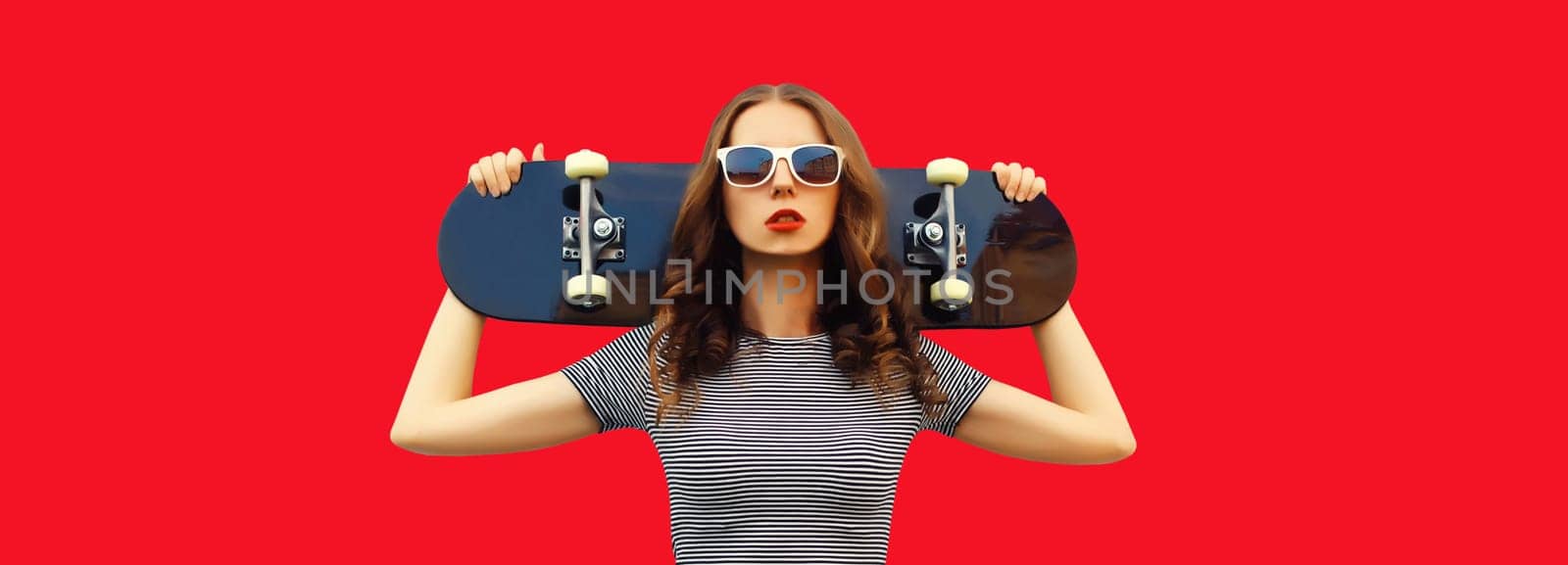 Portrait of stylish young woman posing with skateboard on red studio background