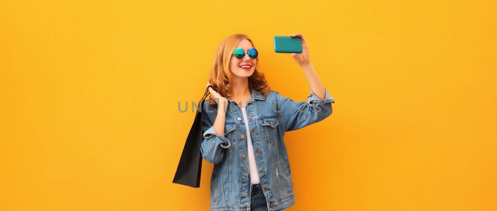 Happy smiling young woman taking selfie with phone holding shopping bags on yellow background by Rohappy