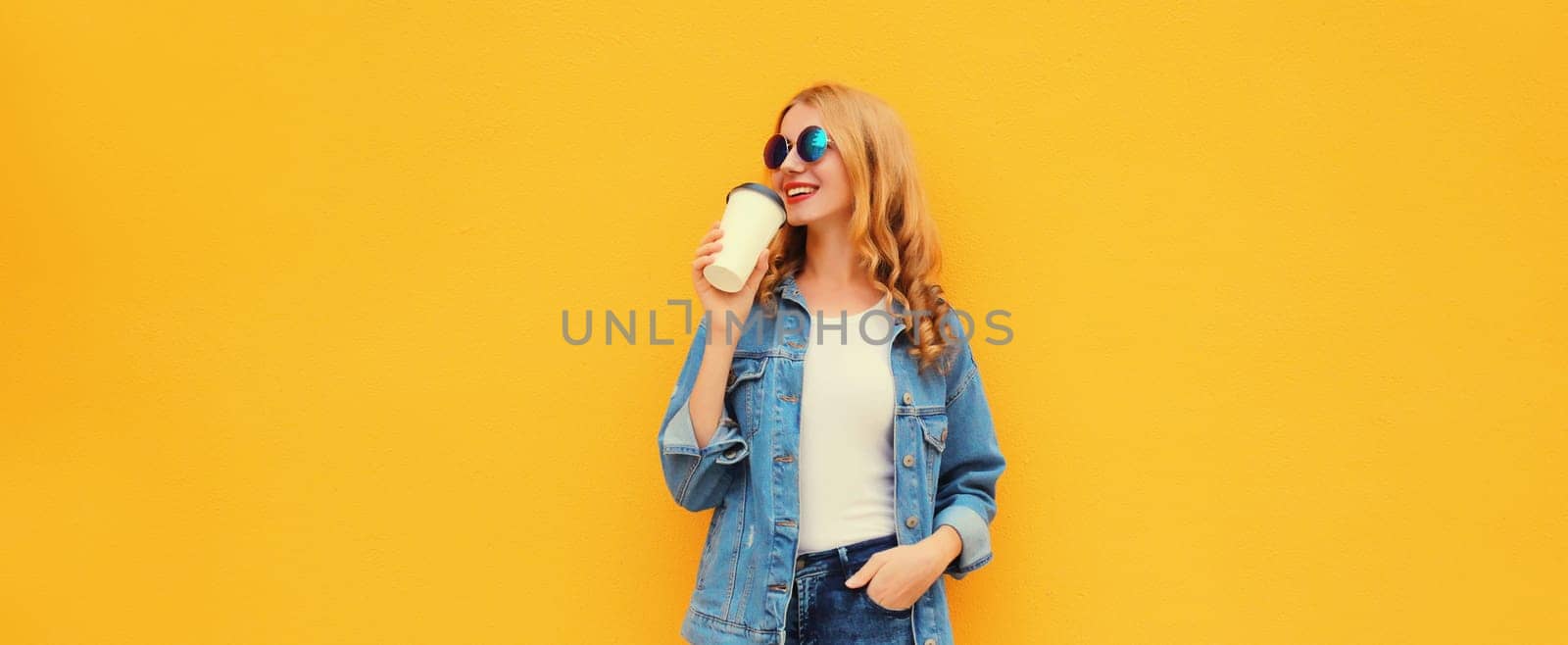 Beautiful happy smiling young woman with cup of coffee in denim jacket on yellow studio background