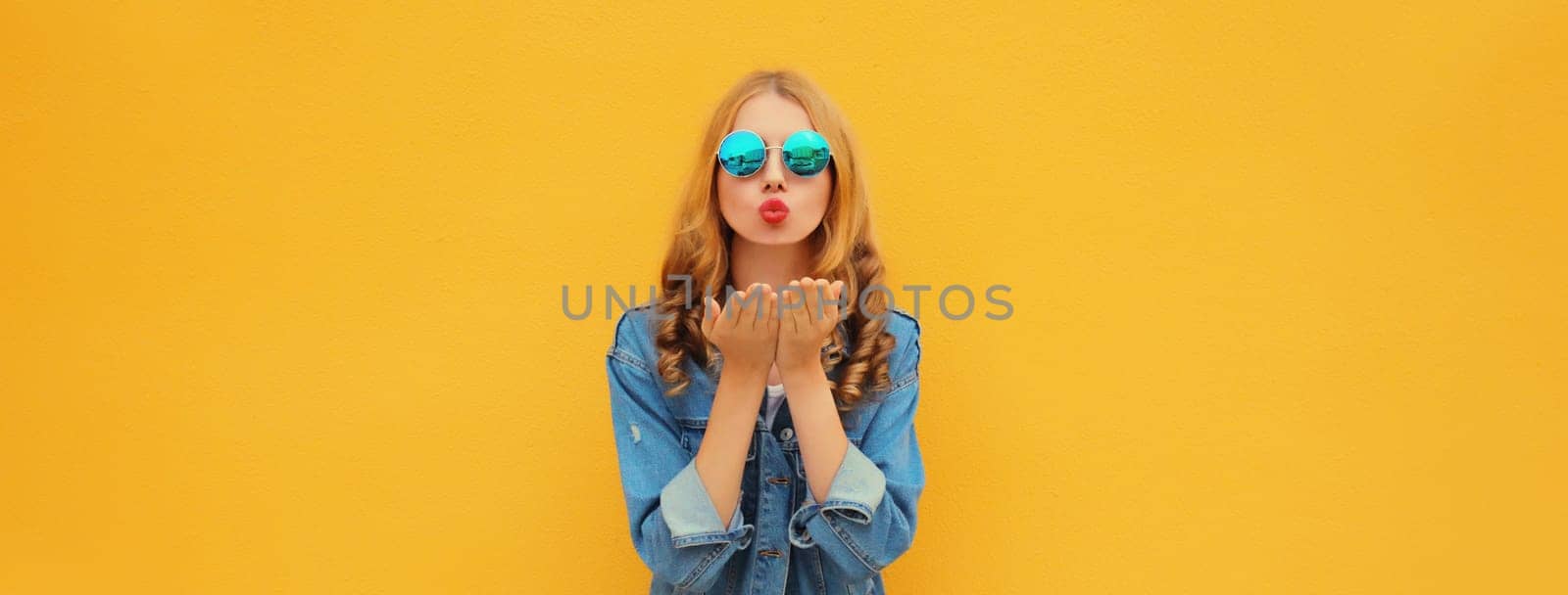Portrait of beautiful stylish young woman blowing kiss in round sunglasses, denim jacket posing on yellow studio background