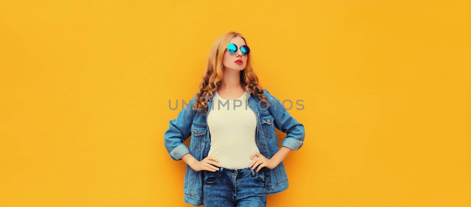 Beautiful modern young blonde woman posing in denim jacket, round sunglasses on yellow studio background