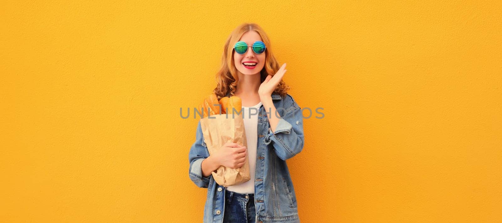 Portrait of happy cheerful smiling young woman holding grocery shopping paper bag with fresh long white bread baguette on yellow studio background