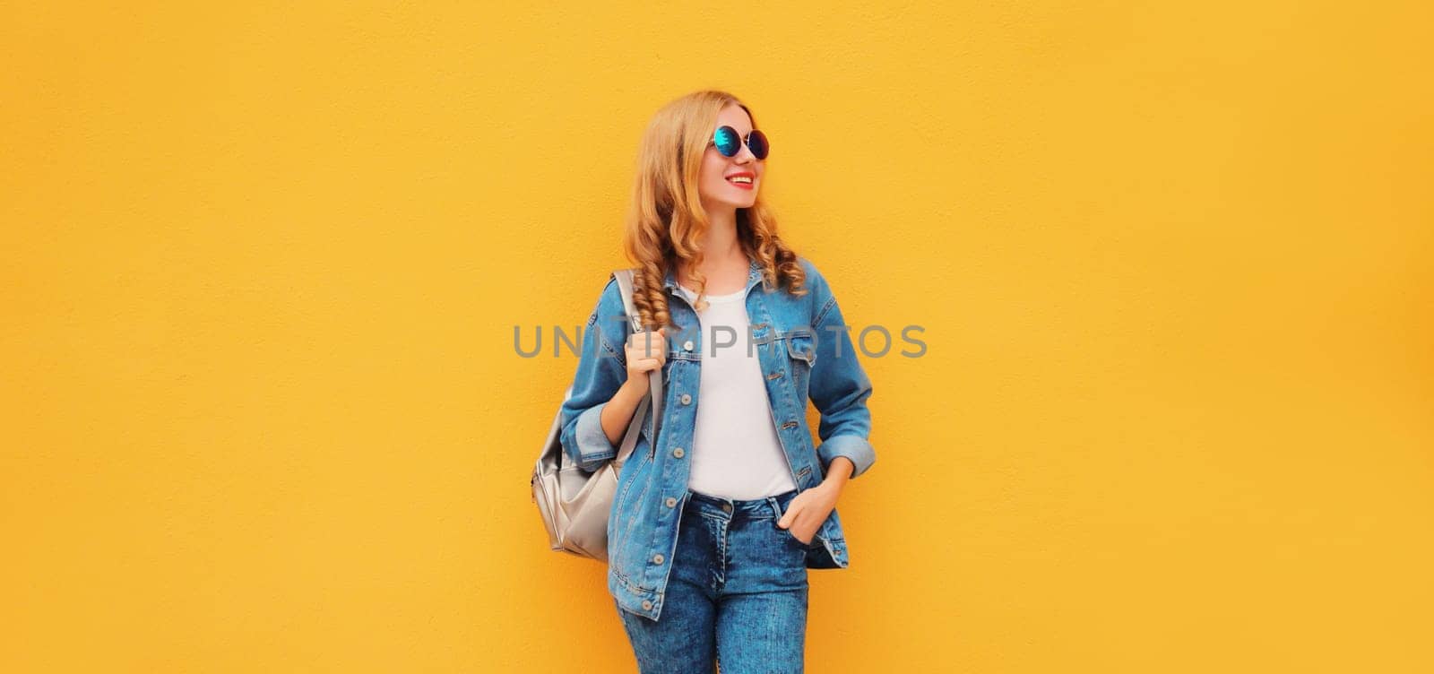 Portrait of stylish modern smiling young woman with backpack in denim jacket on yellow background by Rohappy