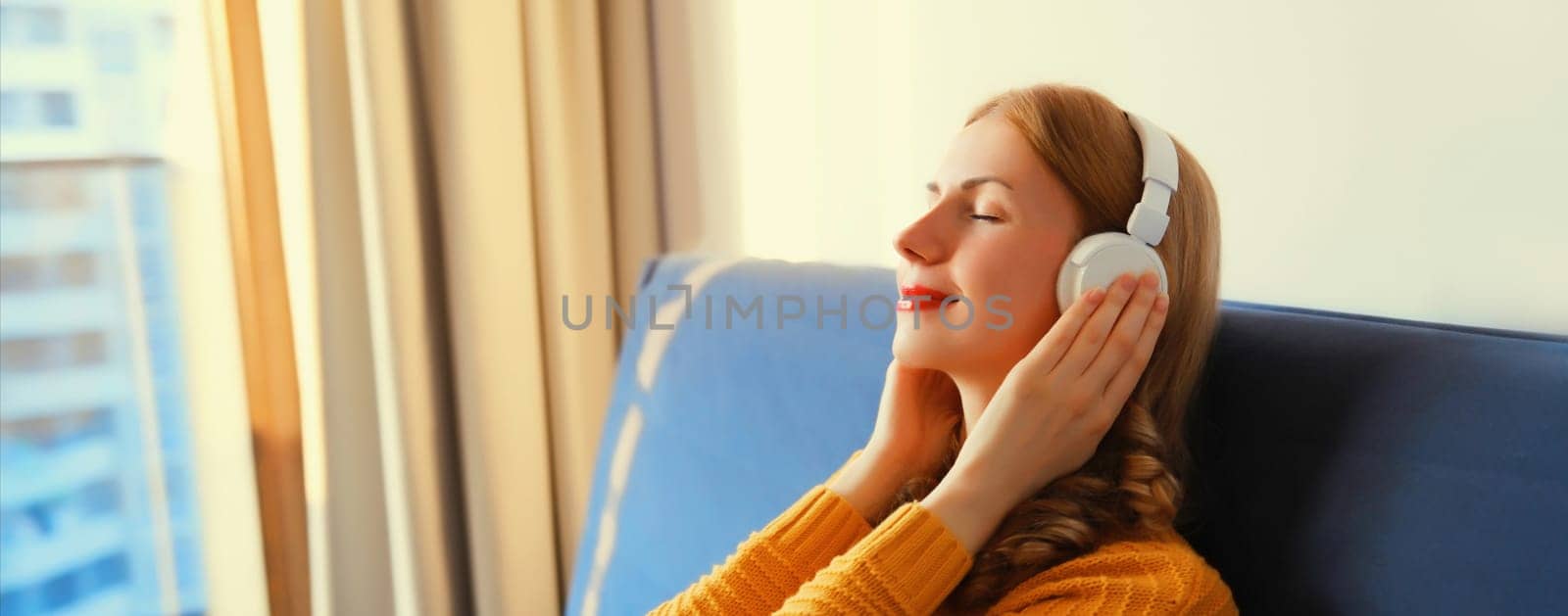 Happy relaxed young woman listening to music with wireless headphones with closed eyes lies on the couch at home
