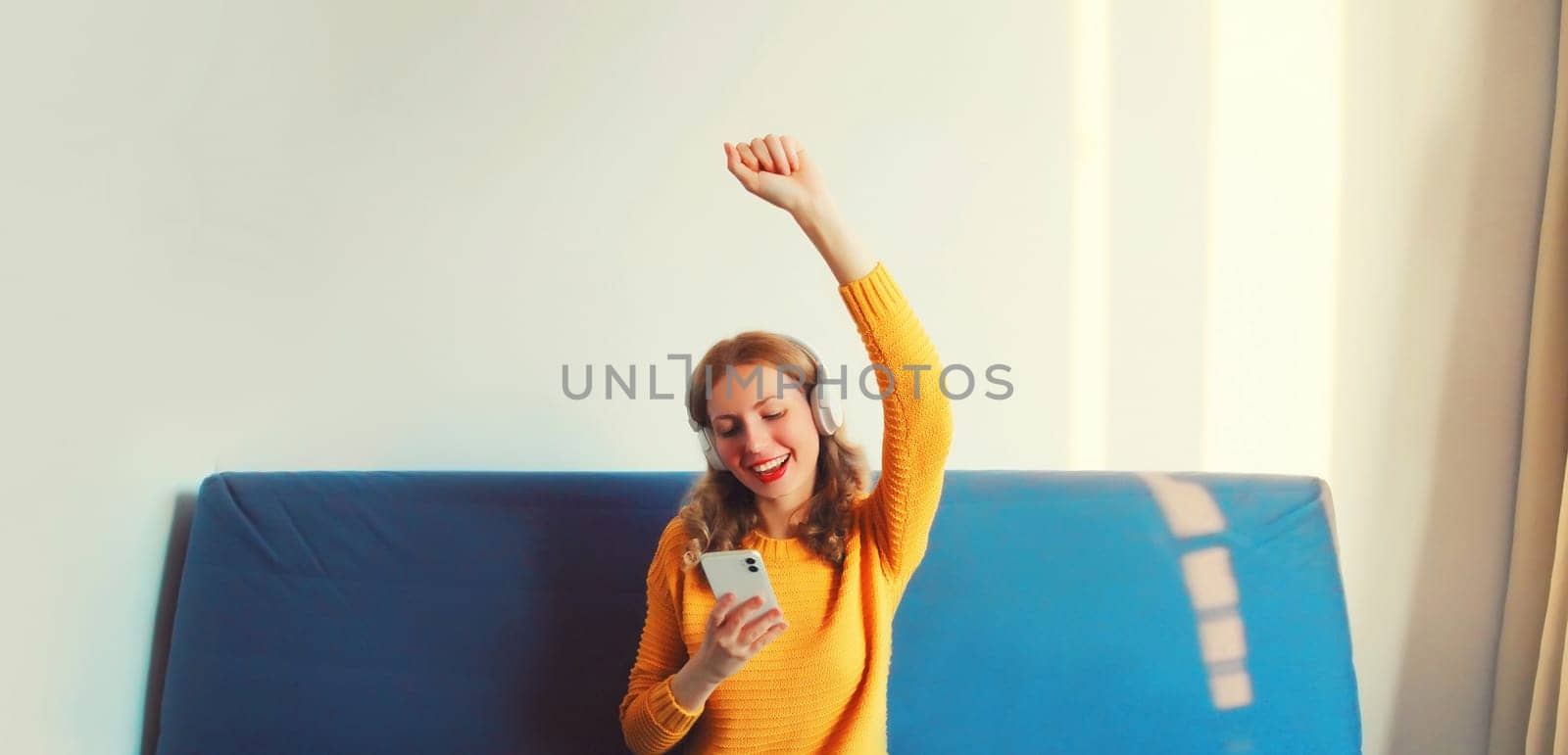 Happy cheerful young woman holding smartphone listening to music with headphones having fun sitting on sofa at home