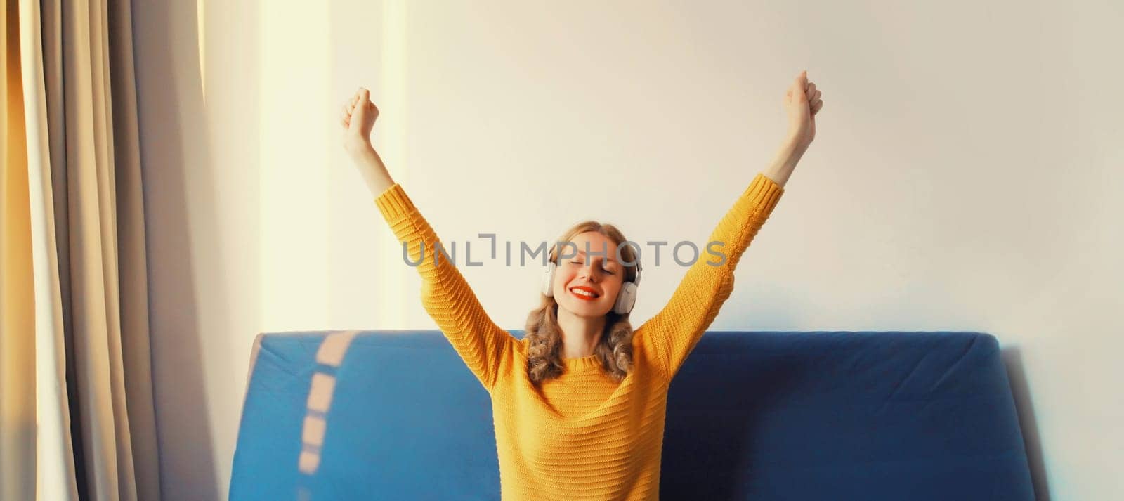 Happy cheerful young woman listening to music with headphones having fun sitting on sofa at home