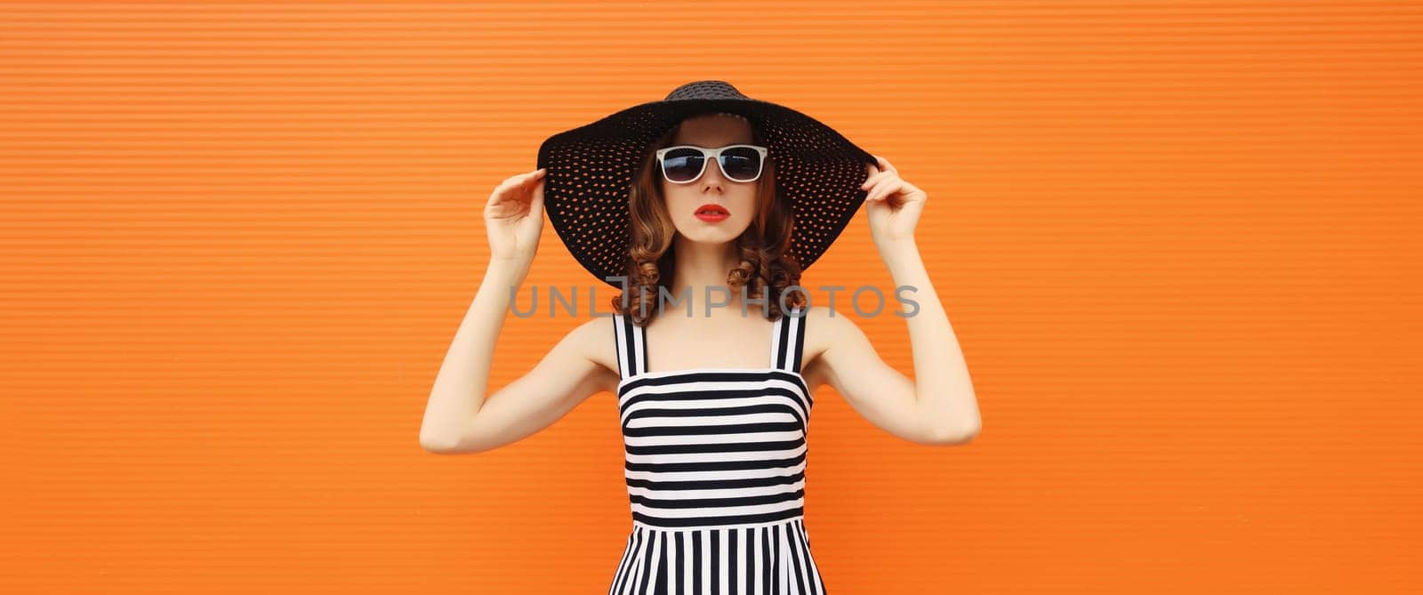 Portrait of beautiful young woman posing wearing black summer straw hat and dress on orange background