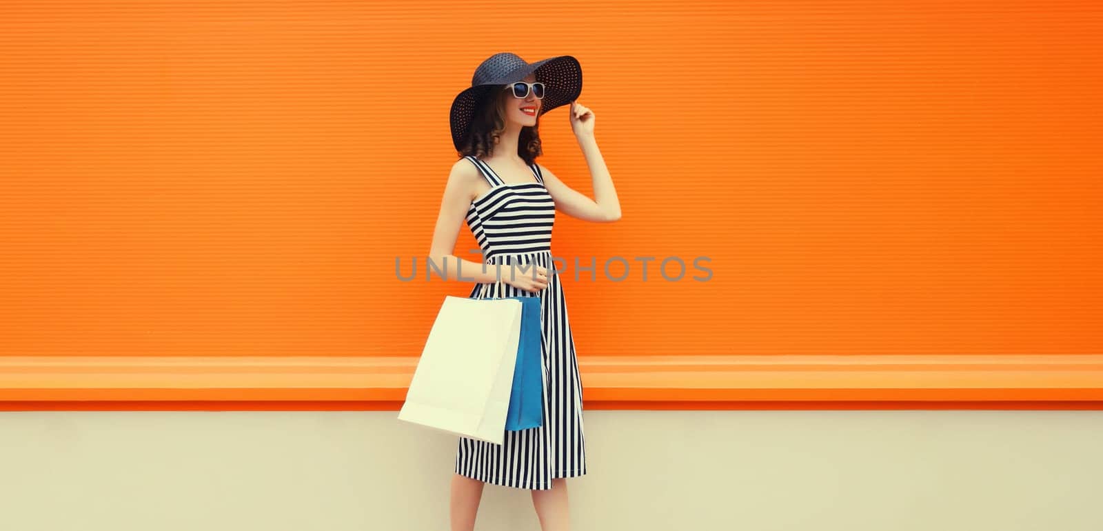 Stylish beautiful happy young woman model with shopping bags in summer black straw hat, striped dress on orange background