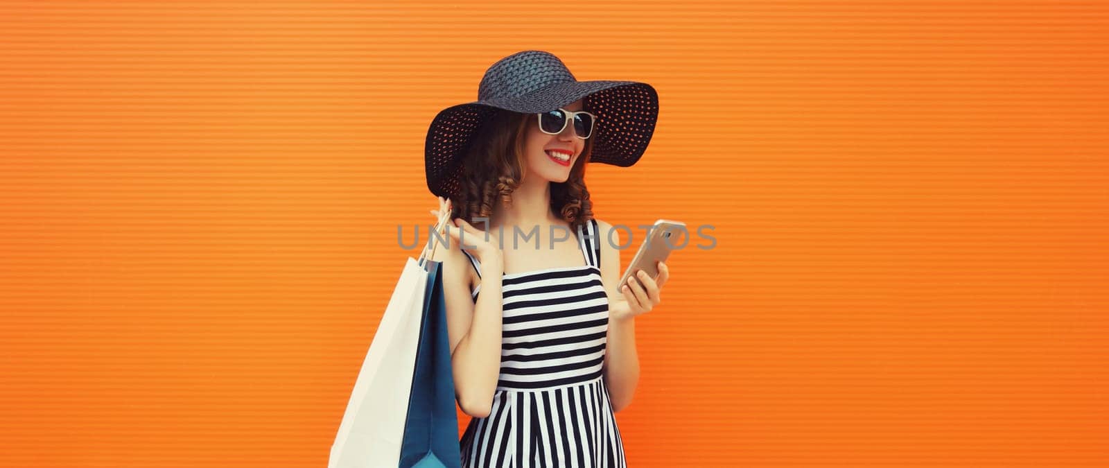 Beautiful happy young woman looking at phone with shopping bag in summer black straw hat, striped dress on orange background