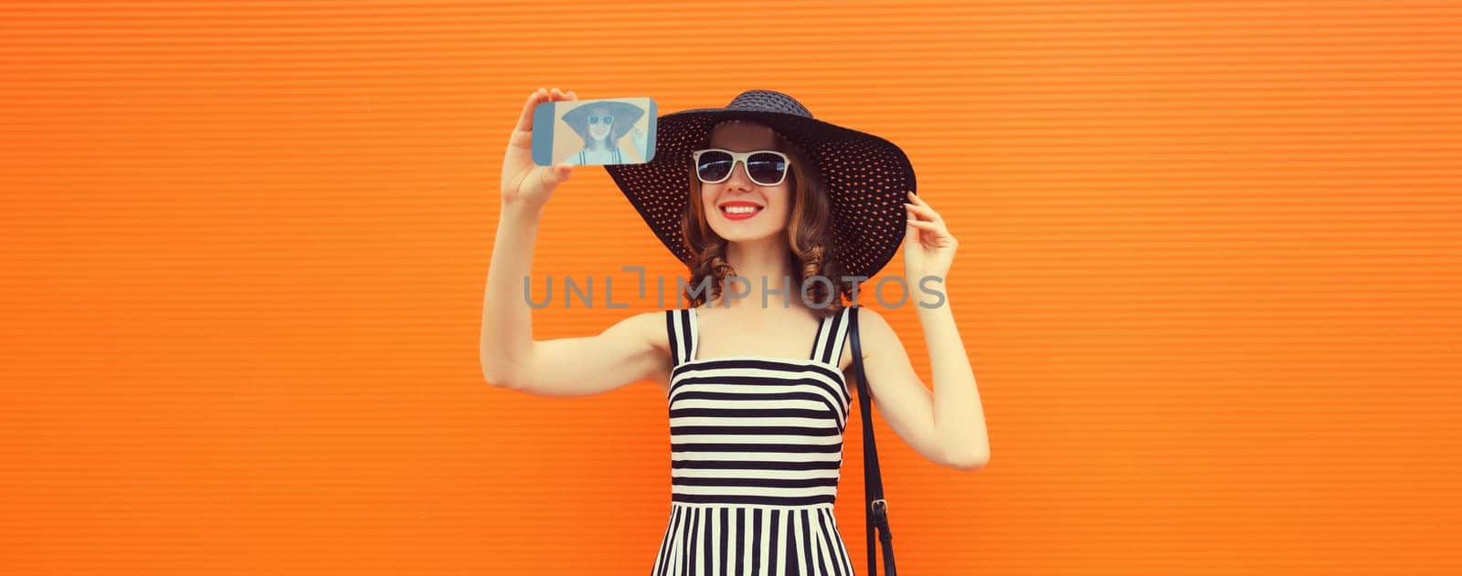 Portrait of stylish happy smiling young woman taking selfie with mobile phone in summer hat, sunglasses on orange background