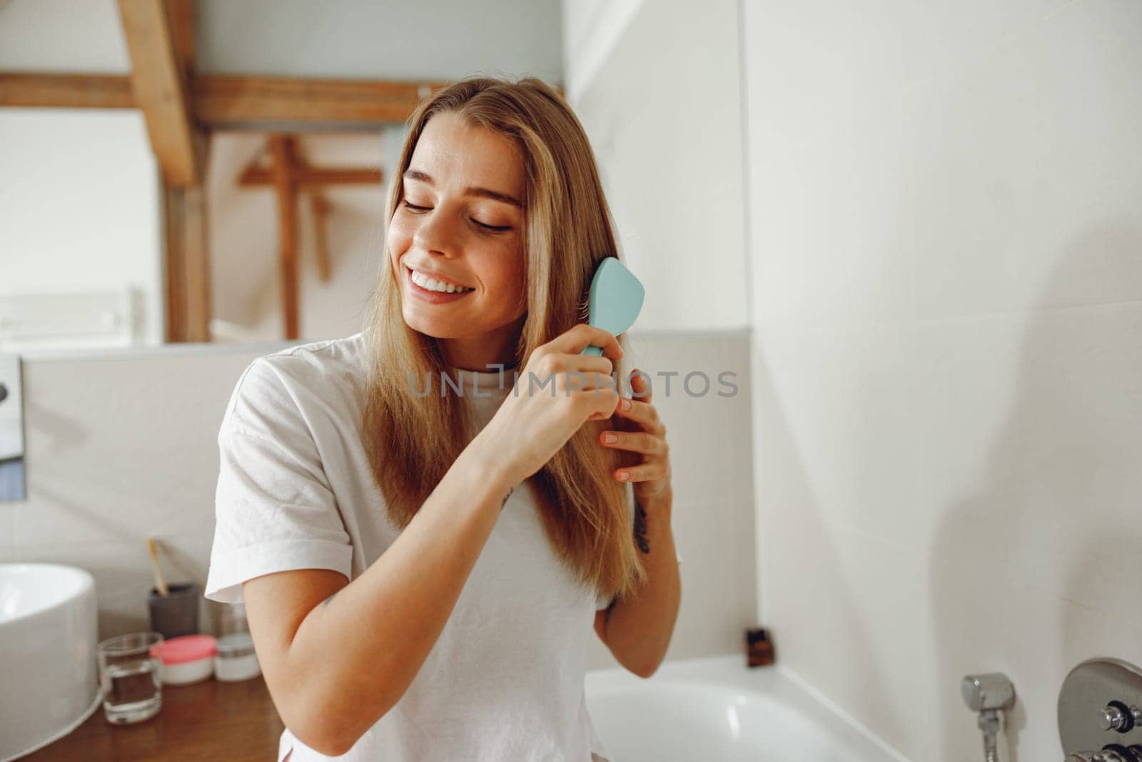 Young woman brushing her hair with comb while standing in bathroom near mirror by Yaroslav_astakhov