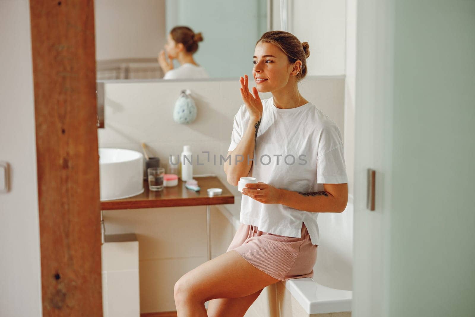 Happy woman applying hydrating moisturizer on her face sitting in bathroom. Home beauty routine by Yaroslav_astakhov