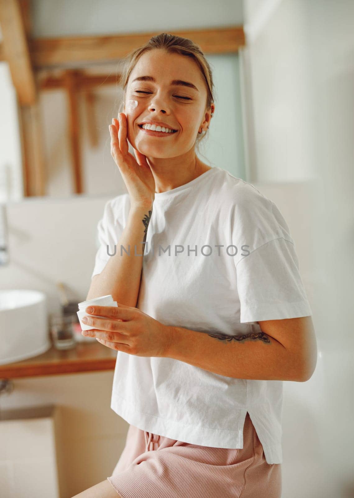 Beautiful young woman taking care of skin by applying moisturizer cream in bathroom