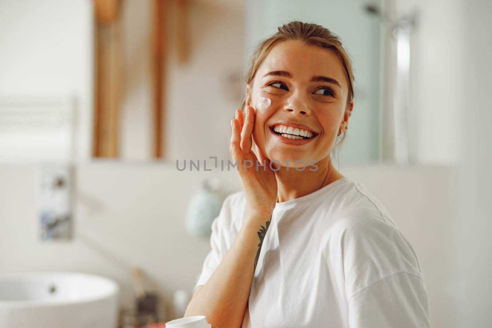 Happy woman applying hydrating moisturizer on her face sitting in bathroom. Home beauty routine