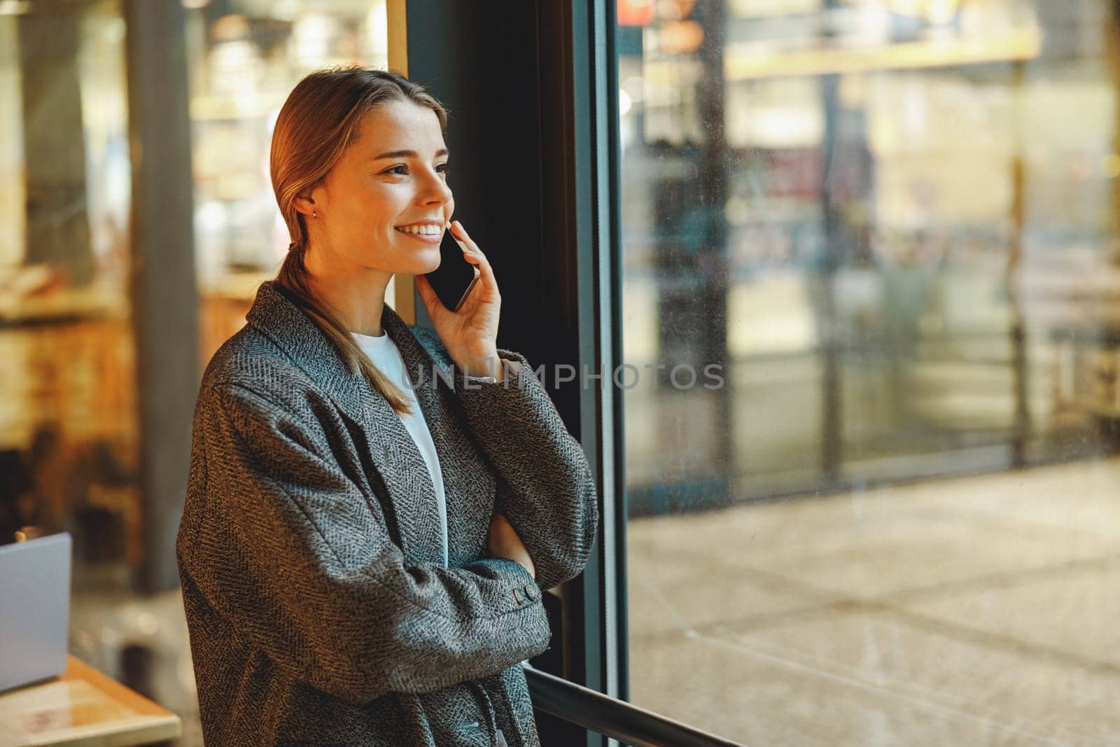 Stylish young female freelancer is talking phone with client while standing in coworking near window