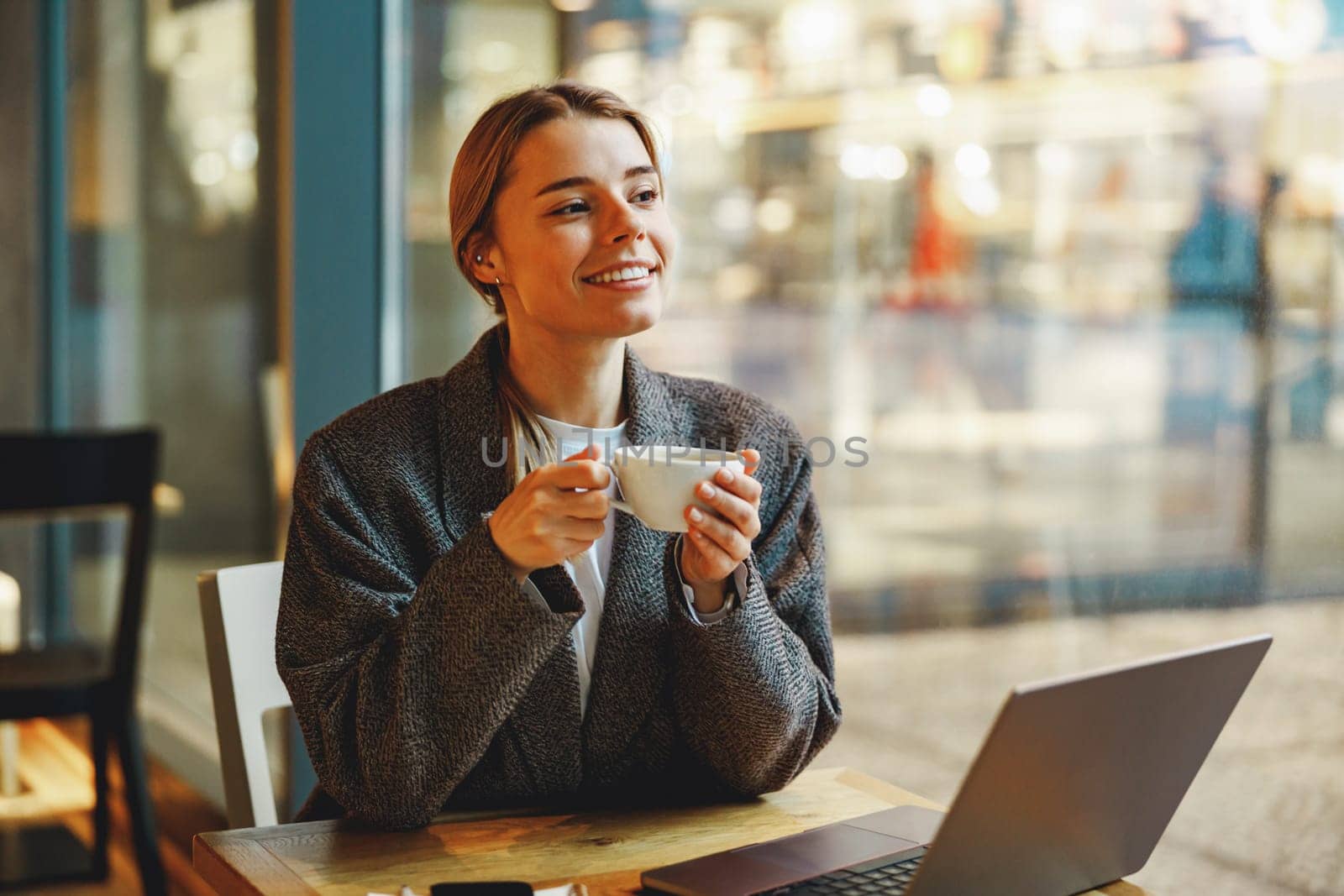 Young female freelancer is drinking coffee in cafe while working on laptop near window by Yaroslav_astakhov