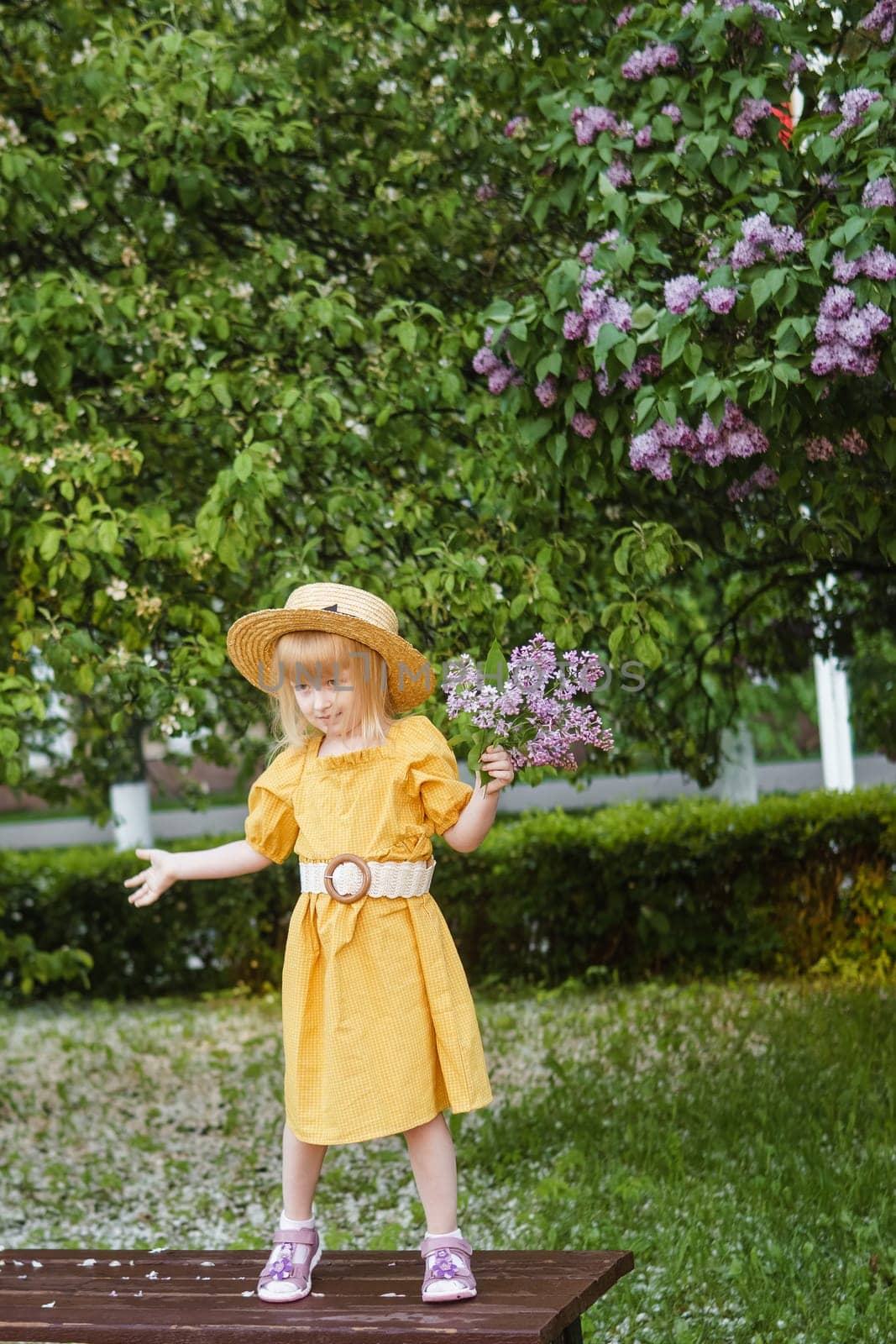 A little girl in a yellow dress and straw hat wearing a bouquet of lilacs. A walk in a spring park, blossoming lilacs