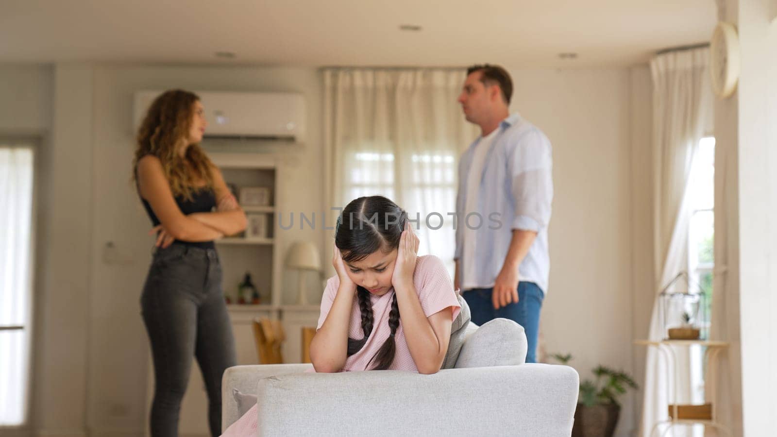 Annoyed and unhappy young girl sitting on sofa trapped in middle of tension by her parent argument in living room. Unhealthy domestic lifestyle and traumatic childhood develop to depression Synchronos